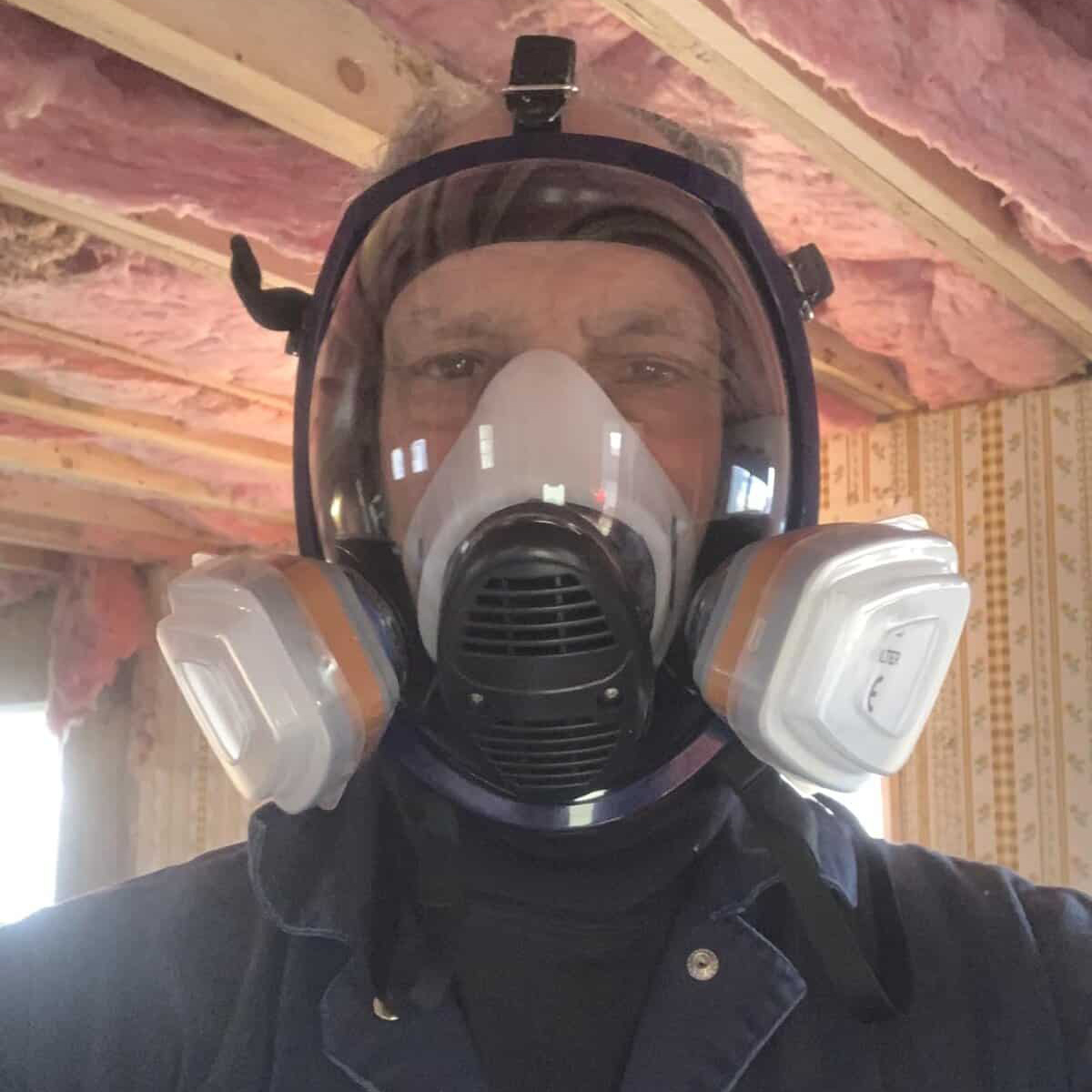 Full-face respirator mask worn by a person during construction or industrial work, with pink insulation and wooden beams in the background.